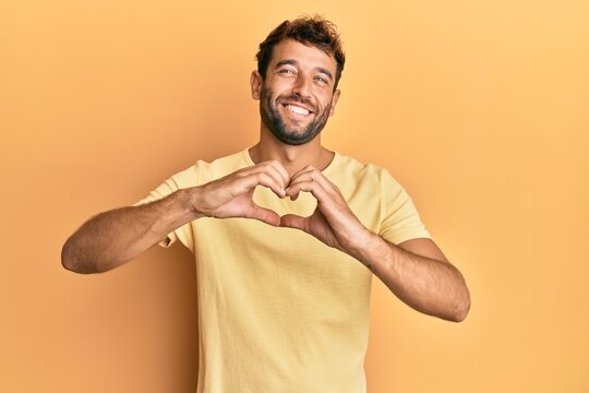 Handsome man with beard wearing casual yellow tshirt over yellow background smiling in love doing heart symbol shape with hands. romantic concept.