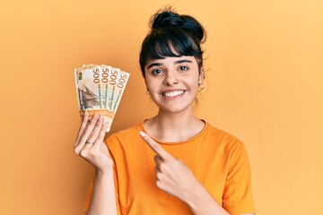 Young hispanic girl holding norwegian krone banknotes smiling happy pointing with hand and finger