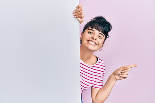 Young hispanic girl holding blank empty banner smiling happy pointing with hand and finger to the side