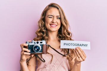 Young blonde girl holding vintage camera and paper with photography word paper winking looking at...