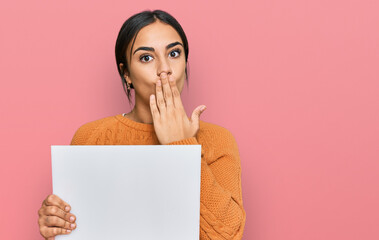 Young brunette woman holding blank empty banner covering mouth with hand, shocked and afraid for...