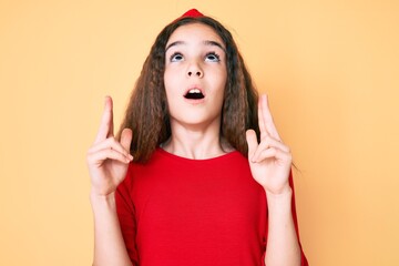 Cute hispanic child girl wearing casual clothes and diadem amazed and surprised looking up and pointing with fingers and raised arms.