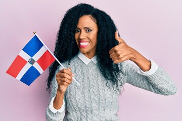 Middle age african american woman holding dominican republic flag smiling happy and positive, thumb up doing excellent and approval sign