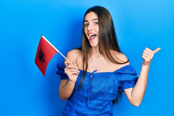 Young brunette teenager holding albania flag pointing thumb up to the side smiling happy with open mouth