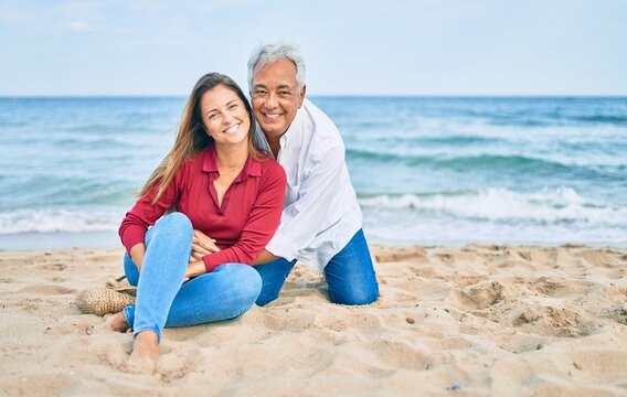 Middle age hispanic couple smiling happy and hugging sitting at the beach