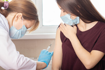Doctor in mask giving an injection of a vaccine to a teenager girl in a medical clinic, vaccine...