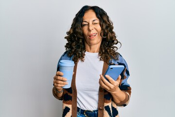 Middle age hispanic woman drinking a cup of coffee and looking at the smartphone screen winking looking at the camera with sexy expression, cheerful and happy face.