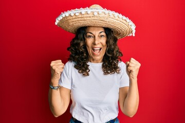 Middle age hispanic woman holding mexican hat screaming proud, celebrating victory and success very excited with raised arms