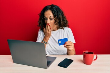 Beautiful middle age woman working at the office using laptop and credit card covering mouth with hand, shocked and afraid for mistake. surprised expression