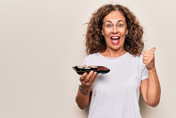 Middle age beautiful woman holding tray with maki sushi over isolated white background pointing thumb up to the side smiling happy with open mouth