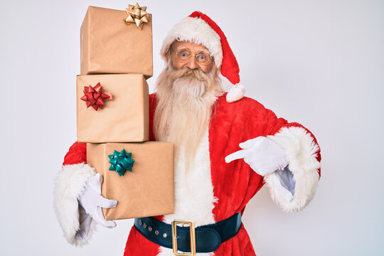 Old senior man with grey hair and long beard wearing santa claus costume holding presents pointing finger to one self smiling happy and proud
