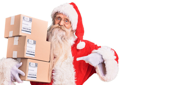Old senior man with grey hair and long beard wearing santa claus costume holding boxes pointing finger to one self smiling happy and proud