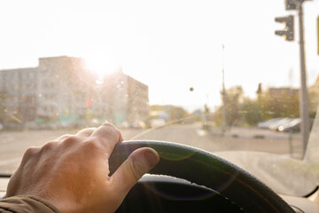 the driver's hand on the steering wheel of a car that is blinded by bright sunlight in the windshield while driving on the road through an intersection with a traffic light