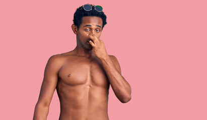 Fototapeta premium African handsome man wearing swimsuit and sunglasses smelling something stinky and disgusting, intolerable smell, holding breath with fingers on nose. bad smell