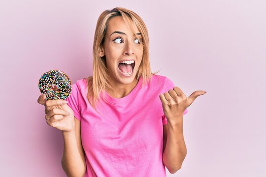 Beautiful blonde woman holding donut pointing thumb up to the side smiling happy with open mouth