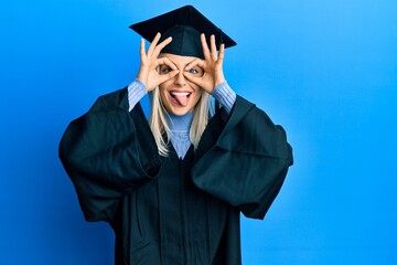 Beautiful blonde woman wearing graduation cap and ceremony robe doing ok gesture like binoculars sticking tongue out, eyes looking through fingers. crazy expression.