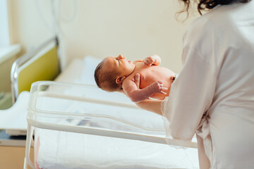 Mother holding hands, calming down, putting her infant baby in hospital bed in postpartum ward.