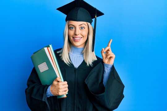 Beautiful blonde woman wearing graduation cap and ceremony robe holding books surprised with an idea or question pointing finger with happy face, number one