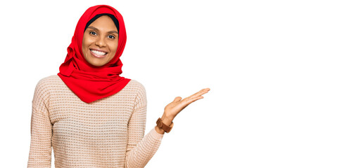 Young african american woman wearing traditional islamic hijab scarf smiling cheerful presenting and pointing with palm of hand looking at the camera.