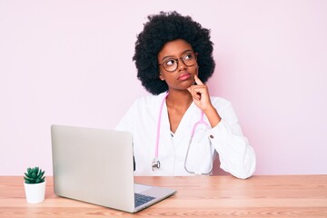 Young african american woman wearing doctor stethoscope working using computer laptop serious face thinking about question with hand on chin, thoughtful about confusing idea