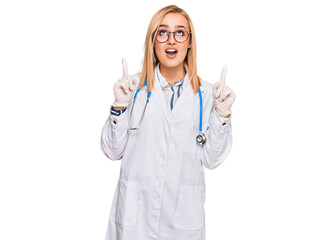 Beautiful caucasian woman wearing doctor uniform and stethoscope amazed and surprised looking up and pointing with fingers and raised arms.