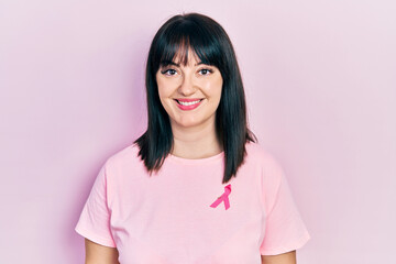 Fototapeta premium Young hispanic woman wearing pink cancer ribbon on shirt looking positive and happy standing and smiling with a confident smile showing teeth