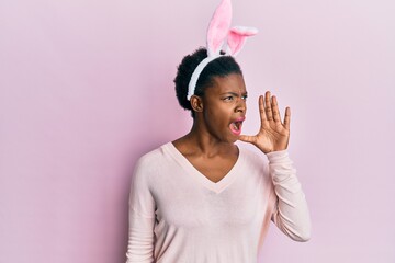 Young african american girl wearing cute easter bunny ears shouting and screaming loud to side with...