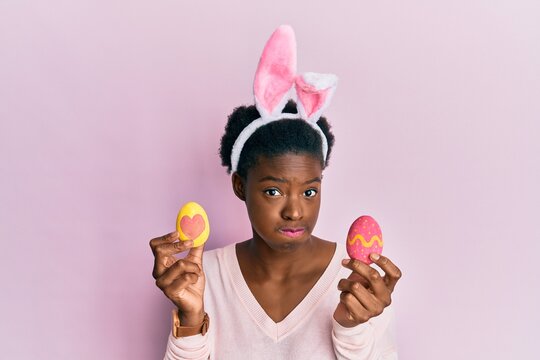 Young african american girl wearing cute easter bunny ears holding painted eggs puffing cheeks with funny face. mouth inflated with air, catching air.
