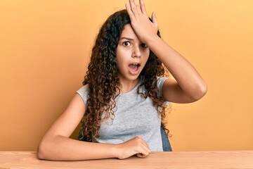 Teenager hispanic girl wearing casual clothes sitting on the table surprised with hand on head for...