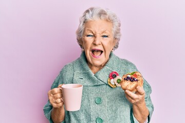 Senior grey-haired woman drinking a cup of coffee and eating bun smiling and laughing hard out loud...