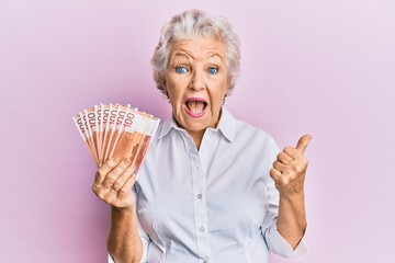 Senior grey-haired woman holding 100 norwegian krone banknotes pointing thumb up to the side...