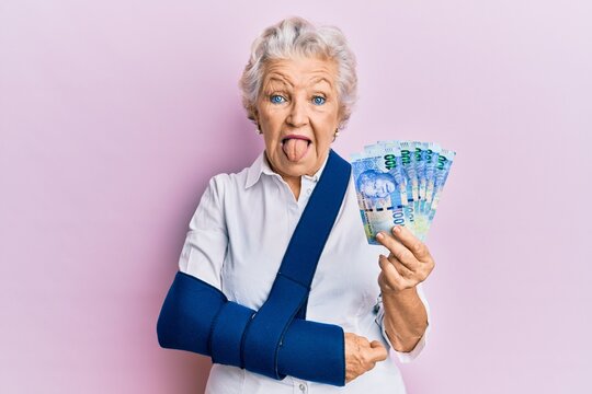 Senior grey-haired woman wearing arm on sling holding south africa rands banknotes sticking tongue out happy with funny expression.