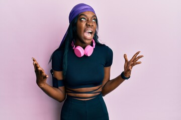 Young african american woman wearing gym clothes and using headphones crazy and mad shouting and...