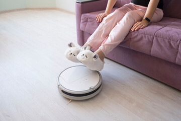 girl in big white sneakers on the couch lifted her feet over the work of a white robot vacuum cleaner