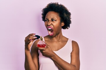 Young african american woman holding perfume angry and mad screaming frustrated and furious,...