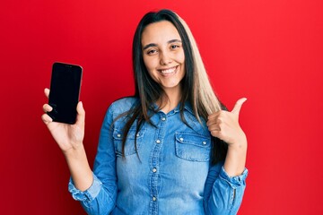 Young hispanic woman holding smartphone showing screen smiling happy and positive, thumb up doing excellent and approval sign