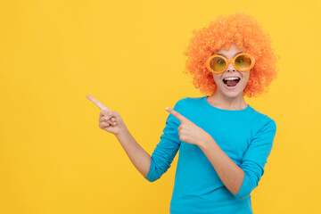 cheerful selfish child girl in curly wig and party glasses pointing finger on copy space, advert