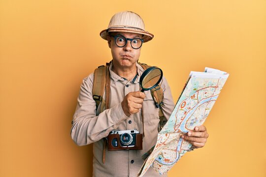 Middle age bald man wearing explorer hat holding magnifying glass on a map puffing cheeks with funny face. mouth inflated with air, catching air.