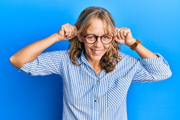 Middle age blonde woman wearing casual clothes and glasses smiling pulling ears with fingers, funny...