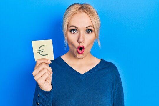 Young blonde woman holding euro symbol reminder scared and amazed with open mouth for surprise, disbelief face