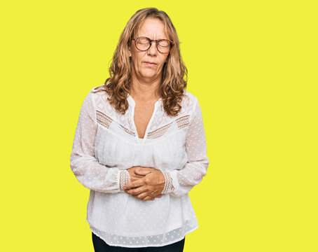 Middle age blonde woman wearing casual white shirt and glasses with hand on stomach because nausea, painful disease feeling unwell. ache concept.