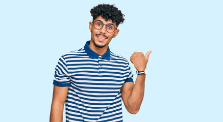 Young arab man wearing casual clothes and glasses smiling with happy face looking and pointing to the side with thumb up.