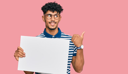 Young arab man holding blank empty banner pointing thumb up to the side smiling happy with open mouth