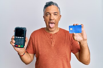 Handsome middle age mature man holding dataphone and credit card sticking tongue out happy with funny expression.