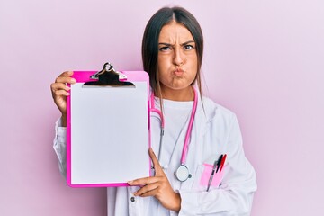 Beautiful hispanic woman wearing doctor stethoscope holding clipboard puffing cheeks with funny...