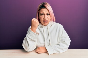 Hispanic woman with pink hair wearing casual sweatshirt sitting on the table angry and mad raising fist frustrated and furious while shouting with anger. rage and aggressive concept.