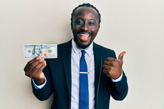 Handsome young black man wearing business suit and tie holding 100 dollars pointing thumb up to the side smiling happy with open mouth