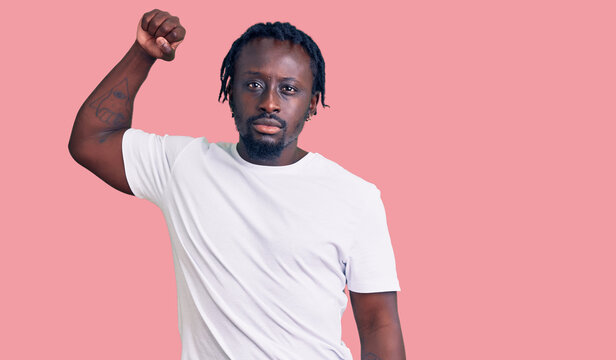 Young african american man with braids wearing casual white tshirt strong person showing arm muscle, confident and proud of power