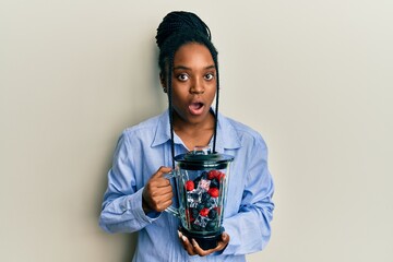 African american woman with braided hair holding food processor mixer machine with fruits afraid and shocked with surprise and amazed expression, fear and excited face.