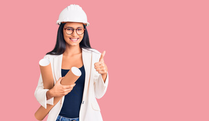 Young beautiful latin girl wearing architect hardhat holding blueprints smiling happy and positive, thumb up doing excellent and approval sign